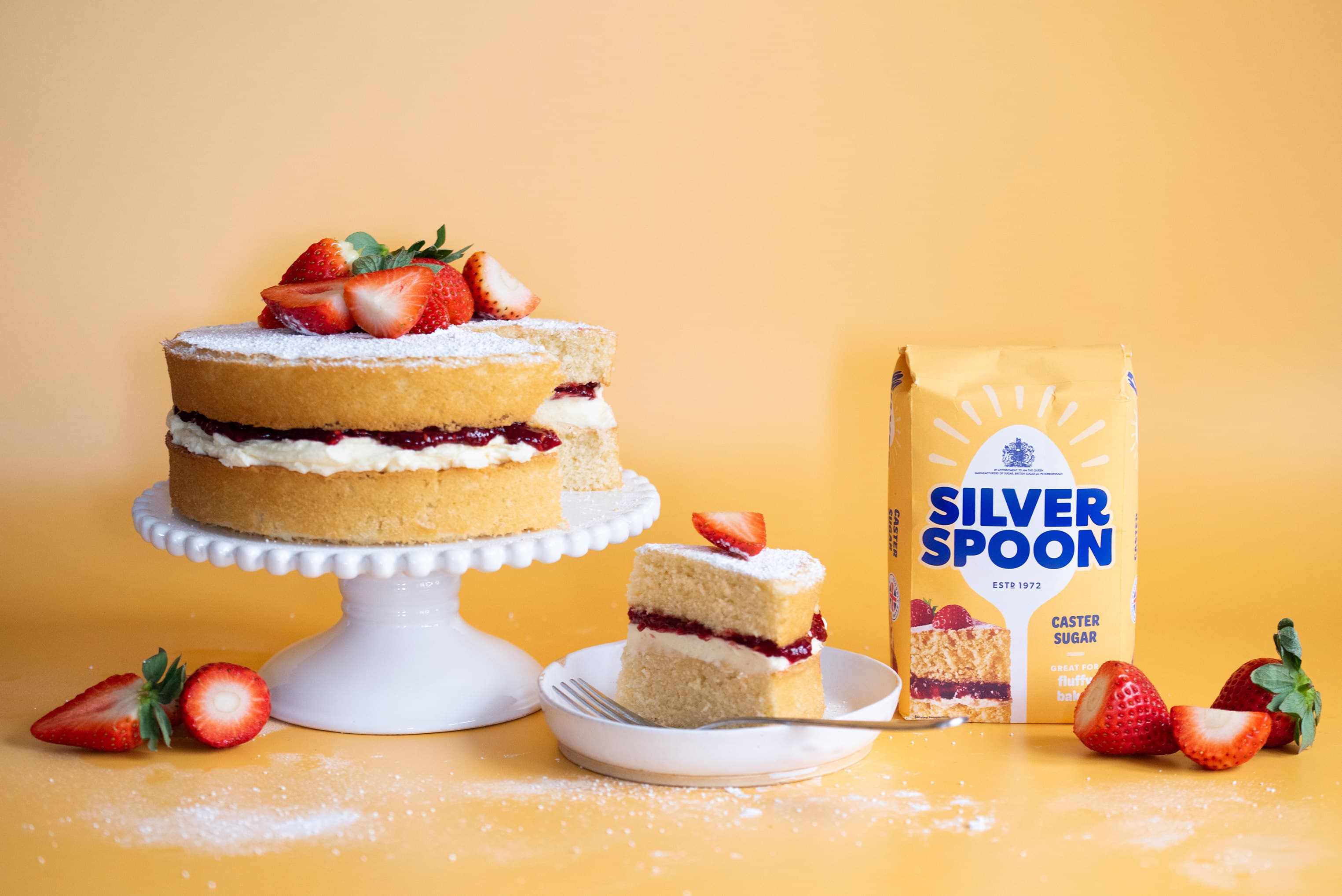 A large tray bake cake, with buttercream topping covered in berries in the style of a Union Jack, alongside packets of Silver Spoon Caster Sugar and Silver Spoon Icing Sugar.