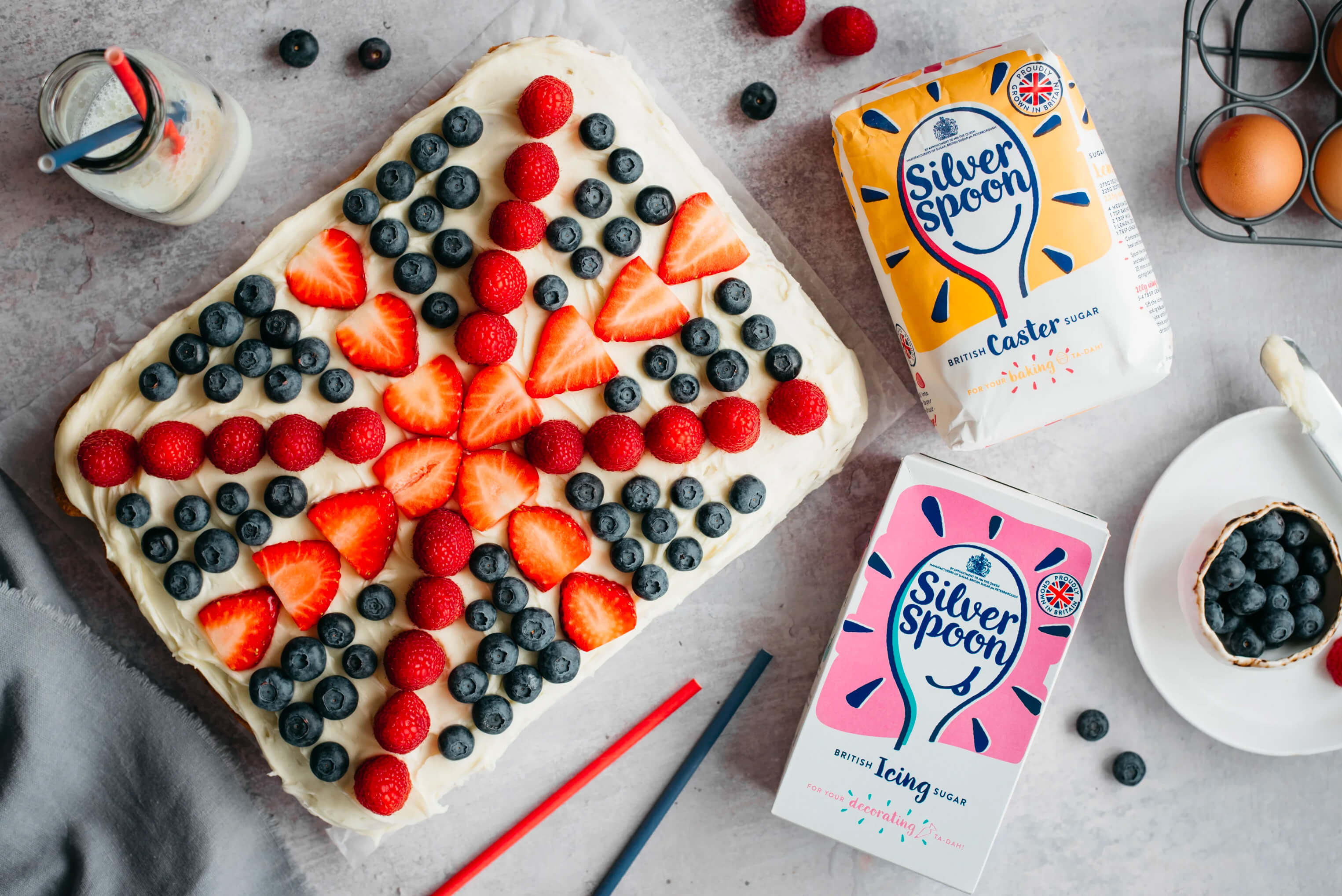 A large tray bake cake, with buttercream topping covered in berries in the style of a Union Jack, alongside packets of Silver Spoon Caster Sugar and Silver Spoon Icing Sugar.